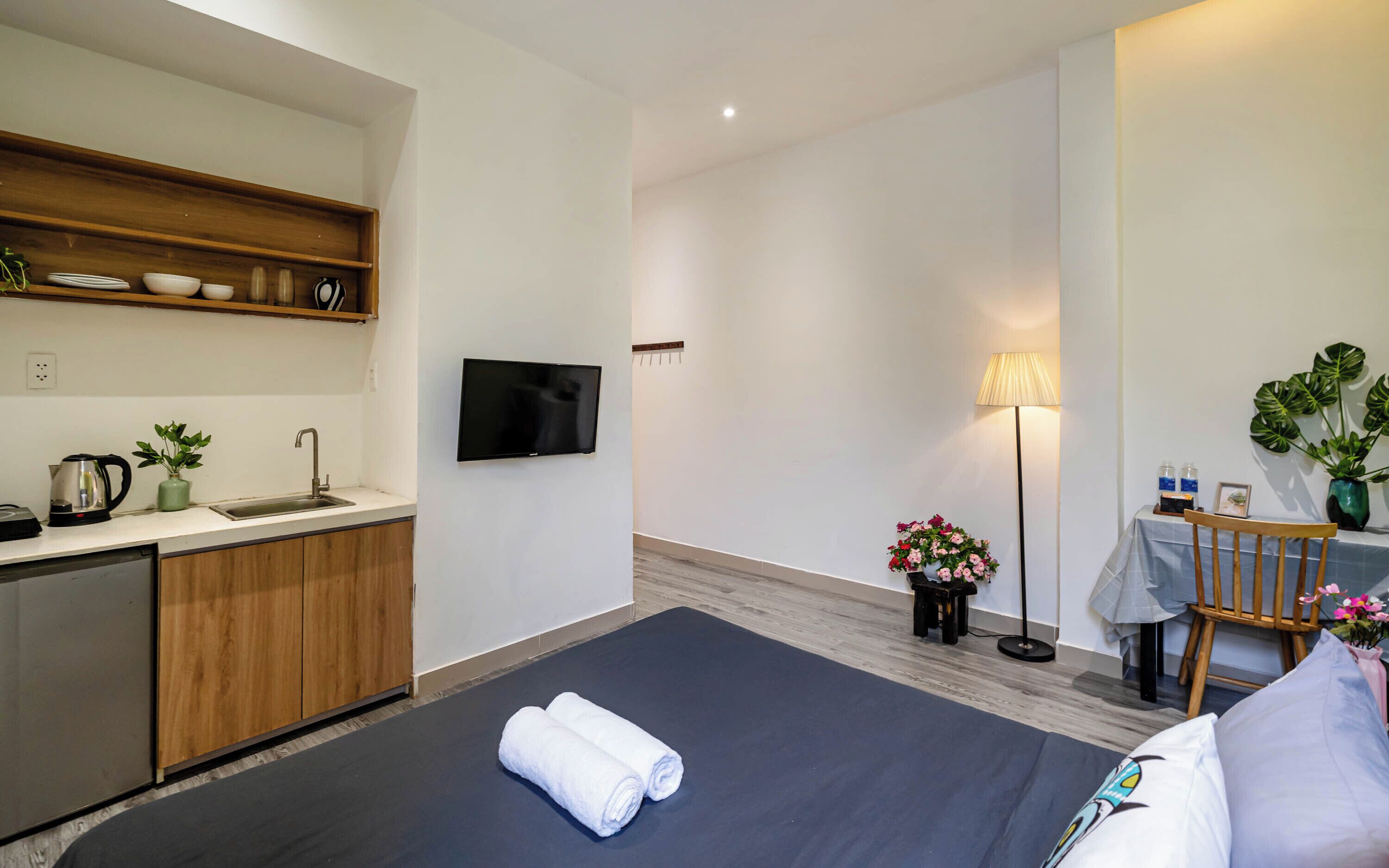 Habitación de hotel con cama, televisión, escritorio con silla, lámpara y una pequeña kitchenette con pileta y placares.