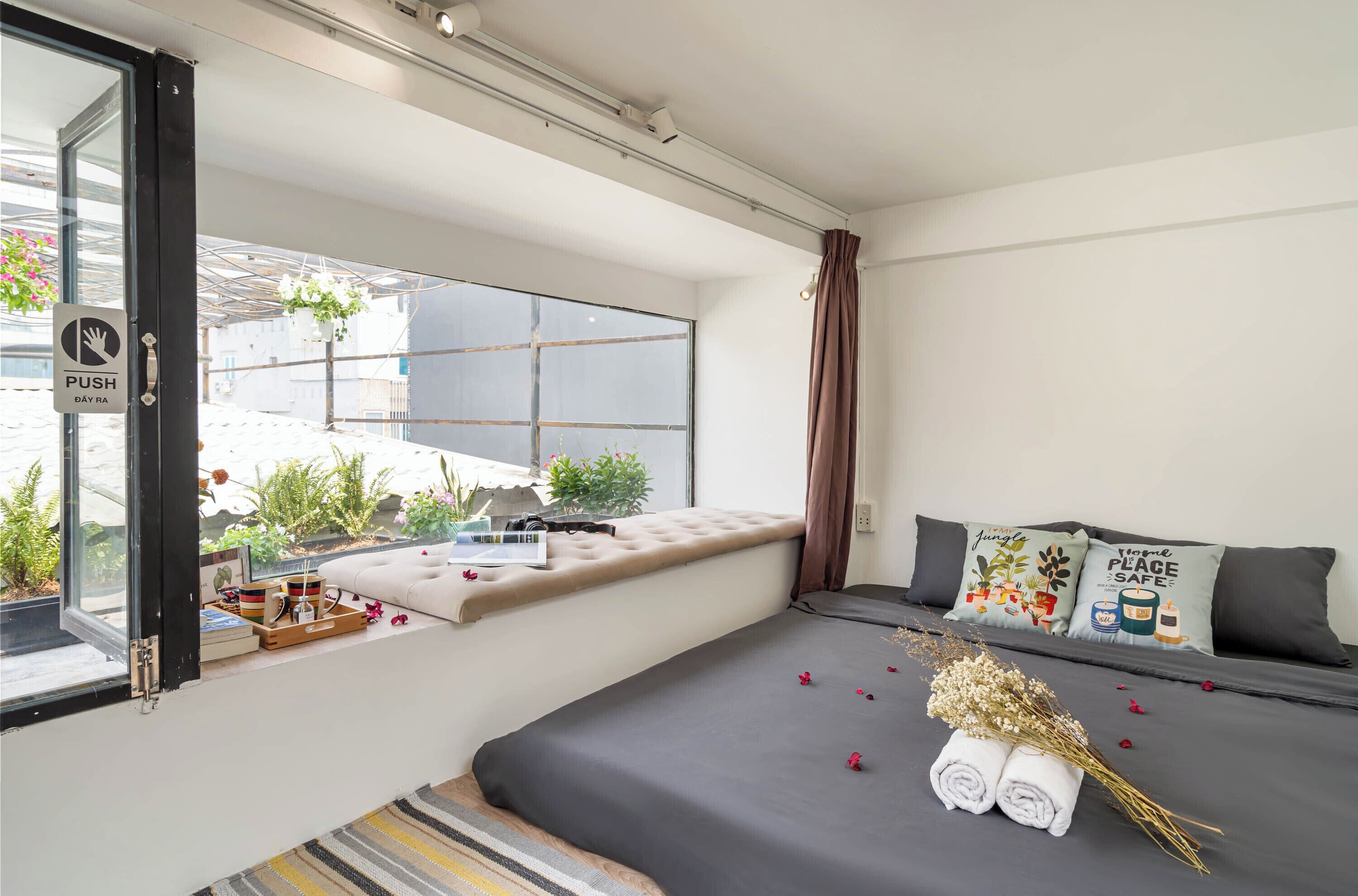 A modern bedroom with a large window, a bed with dark bedding, and a small window seat with a view of greenery.