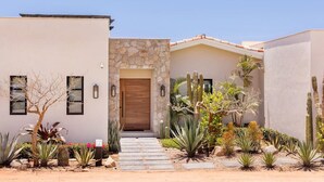 A modern house with a stone facade, a wooden door, and a garden with cacti and desert plants. - Luxurious 4BR Casita – Your Perfect Getaway! (Cabo San Lucas)