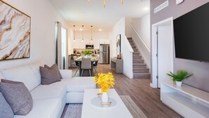 Un salon moderne avec un canapé blanc, une table basse ornée d’un vase de fleurs jaunes, un escalier et une cuisine en arrière-plan.