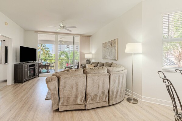 A living room with a large L-shaped sectional sofa, a flat-screen TV, a ceiling fan, and a floor lamp. - Regatta -Resort Style: 3BR/2BA -Lanai w/dining/conversational seating/pool view (Naples)
