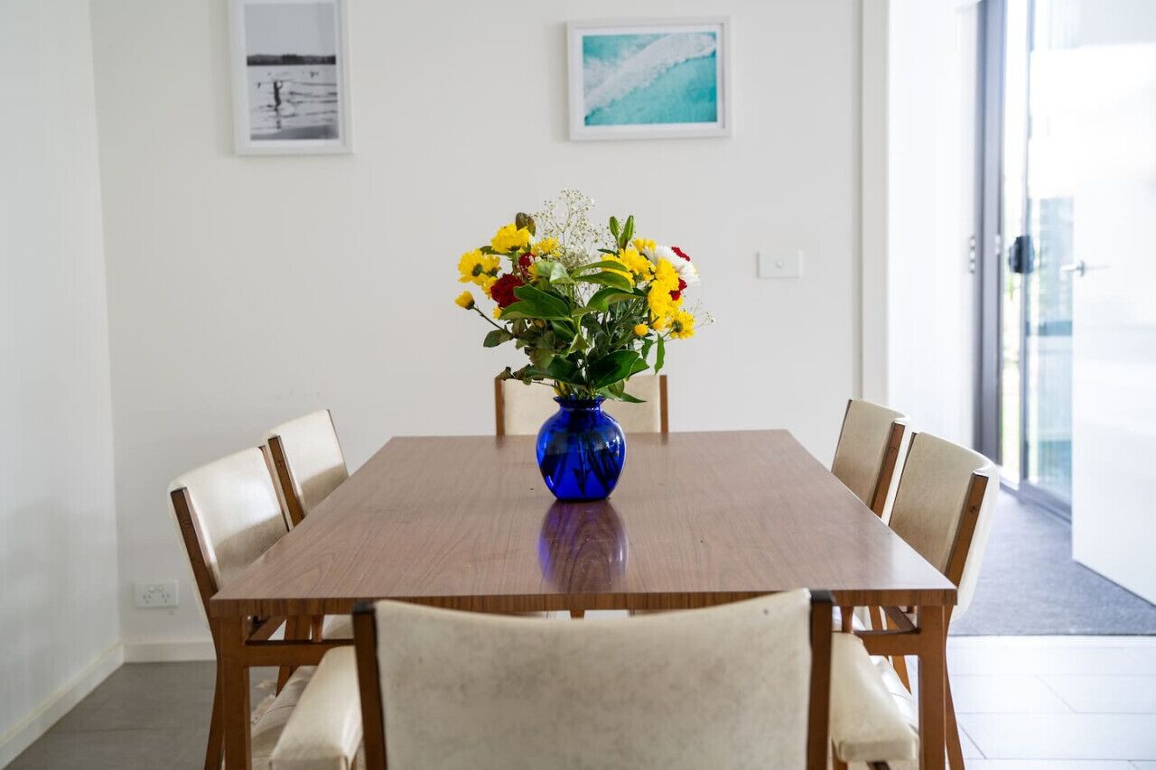A dining area with a wooden table and six chairs, a blue vase with flowers, and two framed pictures on the wall.