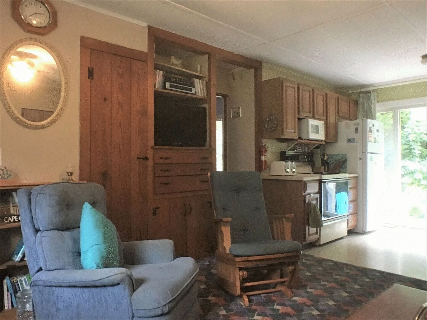 A living room with two armchairs, a wooden cabinet, and a kitchen area with appliances.