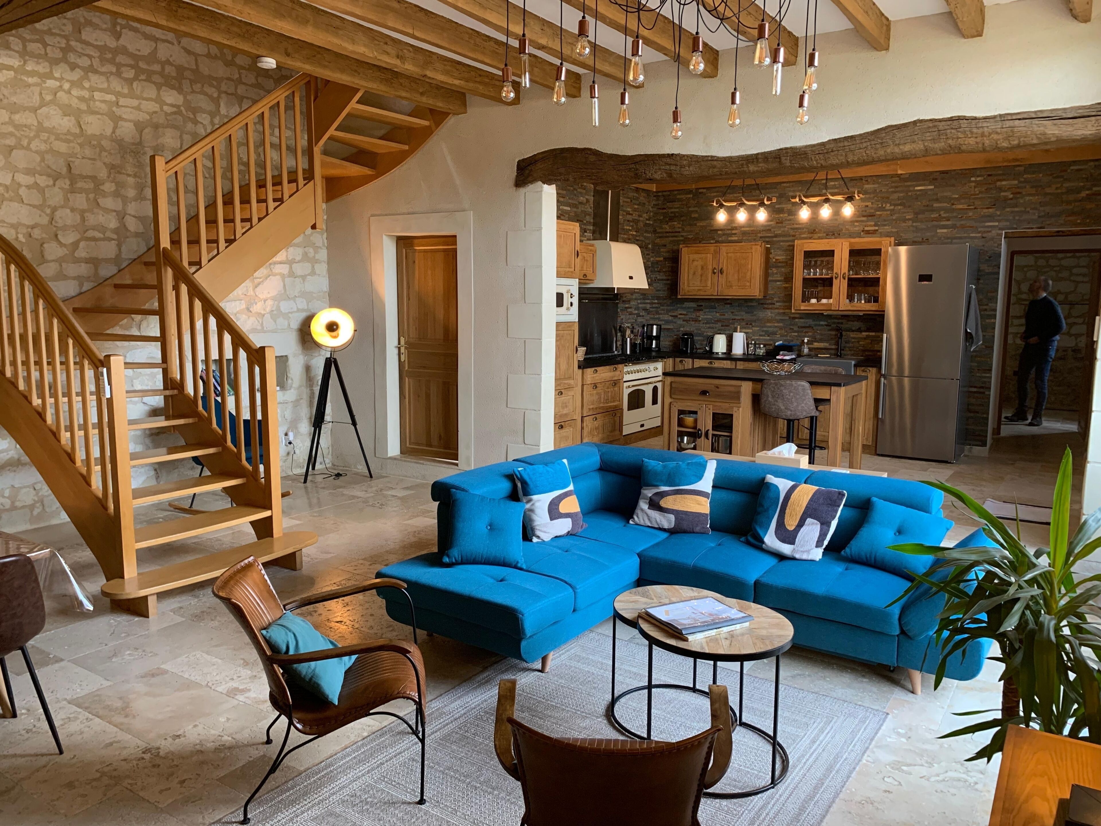 A modern living room with a blue sectional sofa, wooden staircase, and a kitchen area with stone walls and wooden cabinets.