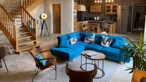 A modern living room with a blue sectional sofa, wooden staircase, and a kitchen area with stone walls and wooden cabinets.