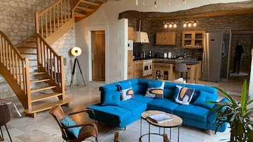 A modern living room with a blue sectional sofa, wooden staircase, and a kitchen area with stone walls and wooden cabinets.