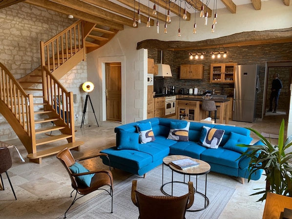 Un salon moderne avec un canapé d’angle bleu, un escalier en bois et un coin cuisine doté de murs en pierre et d’armoires en bois.