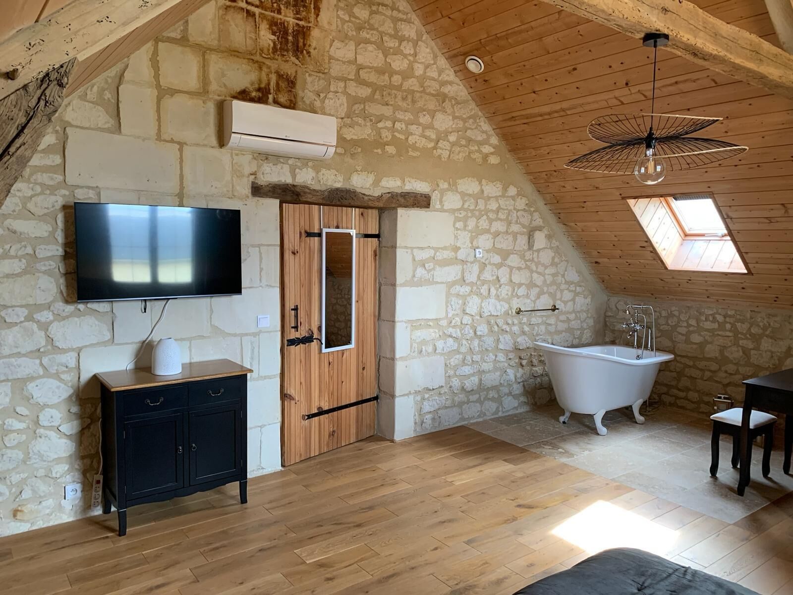 A rustic room with a stone wall, wooden ceiling, a flat-screen TV, a wooden cabinet, a white bathtub, and a skylight.