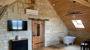 A rustic room with a stone wall, wooden ceiling, a flat-screen TV, a wooden cabinet, a white bathtub, and a skylight.