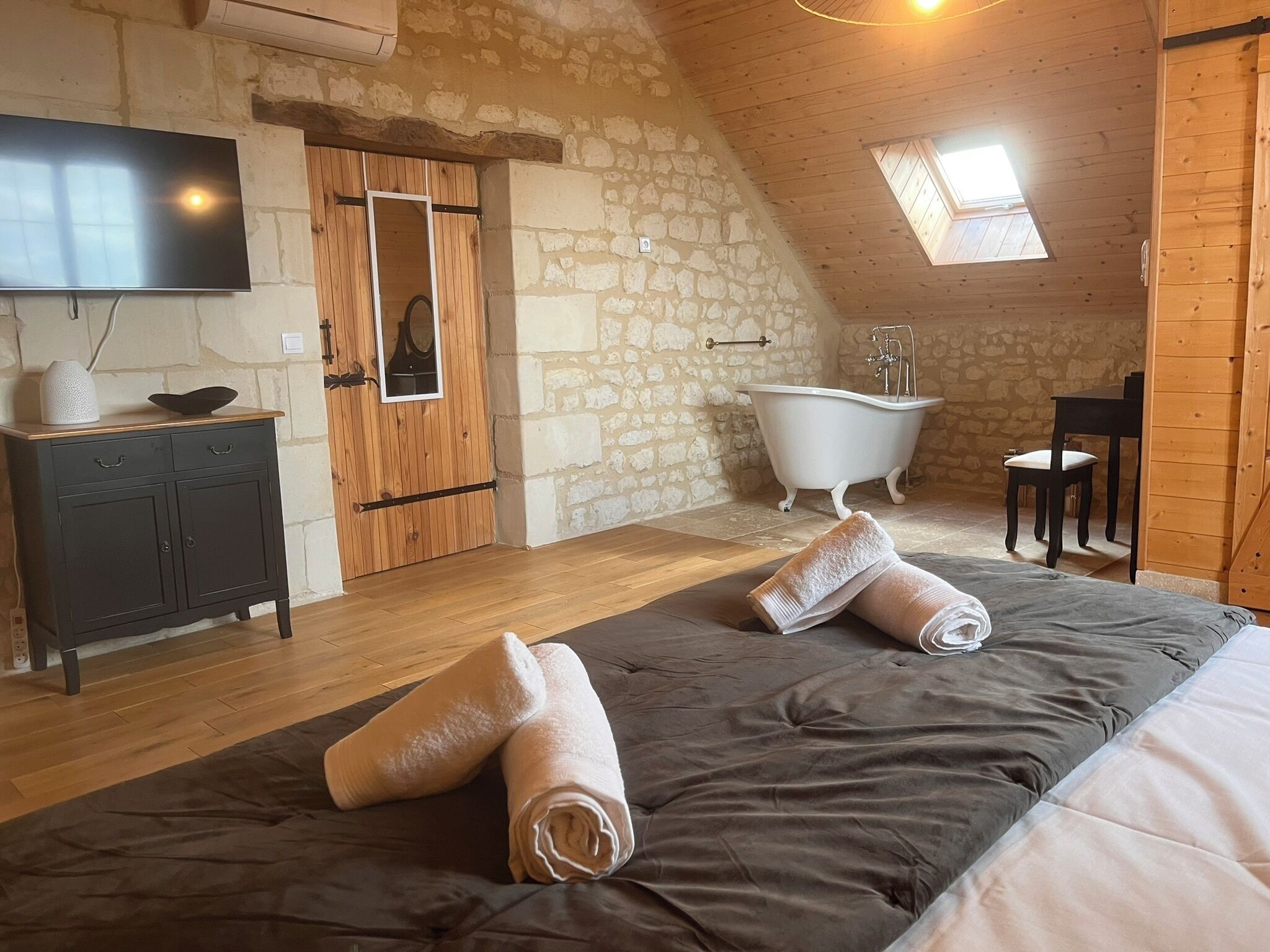A cozy bedroom with a large bed, a wooden dresser, a television, a stone wall, a wooden ceiling, and a skylight.
