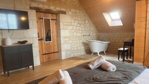 A cozy bedroom with a large bed, a wooden dresser, a television, a stone wall, a wooden ceiling, and a skylight.