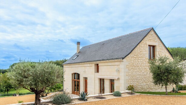 A stone house with a sloped roof, large windows, and a well-maintained garden with various plants and a gravel driveway.