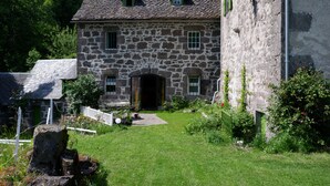 Ein Gebäude aus Stein mit einem geneigten Dach, einem zentralen Eingang und einem kleinen Garten mit weißem Zaun und viel Grün.