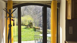 Eine offene Tür führt zu einer Terrasse mit Tisch und Stühlen, einem Kleiderständer und Blick auf eine grüne Fläche mit Bäumen.