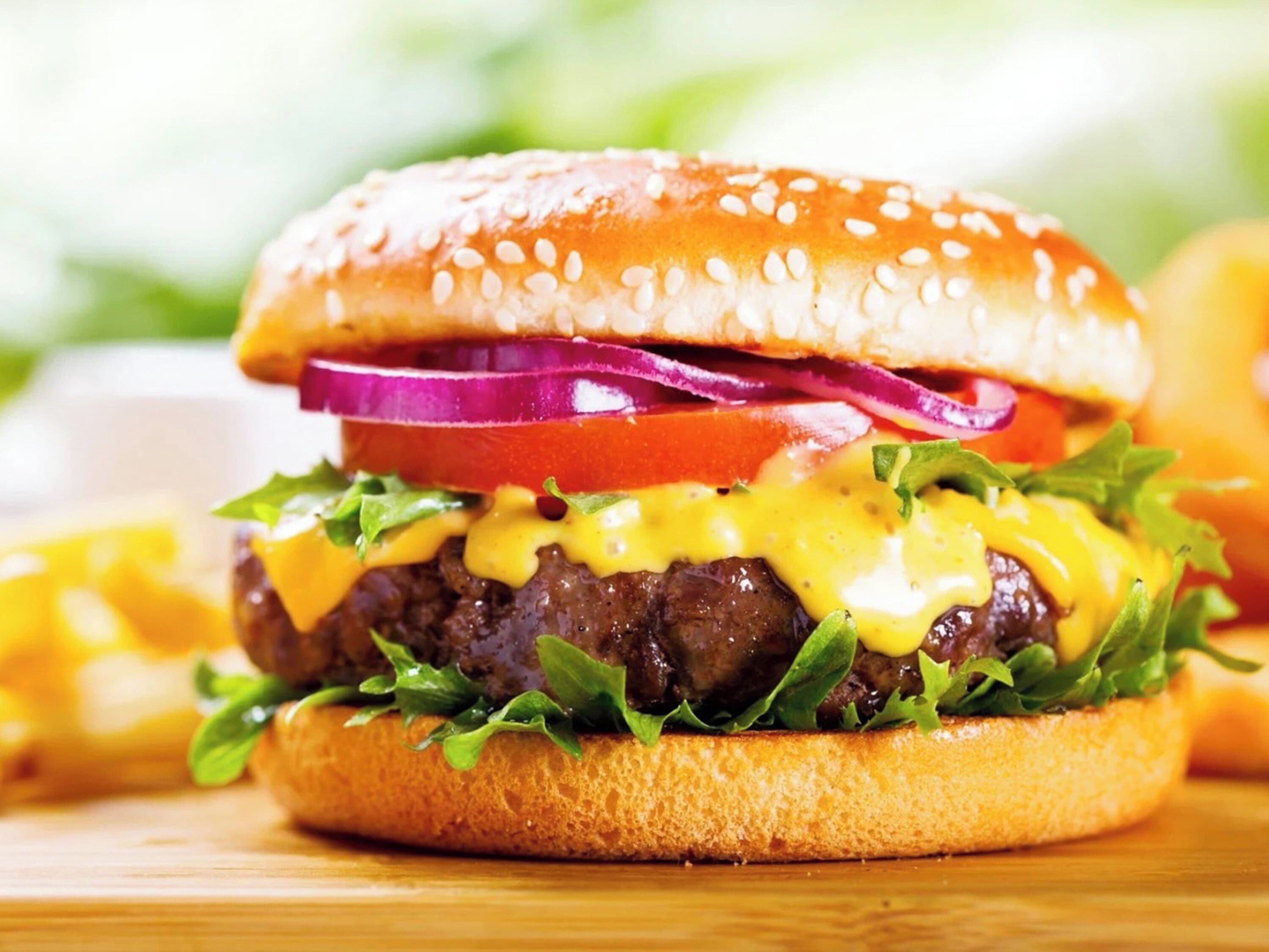 A close-up of a burger with a sesame seed bun, lettuce, tomato, red onion, and melted cheese.