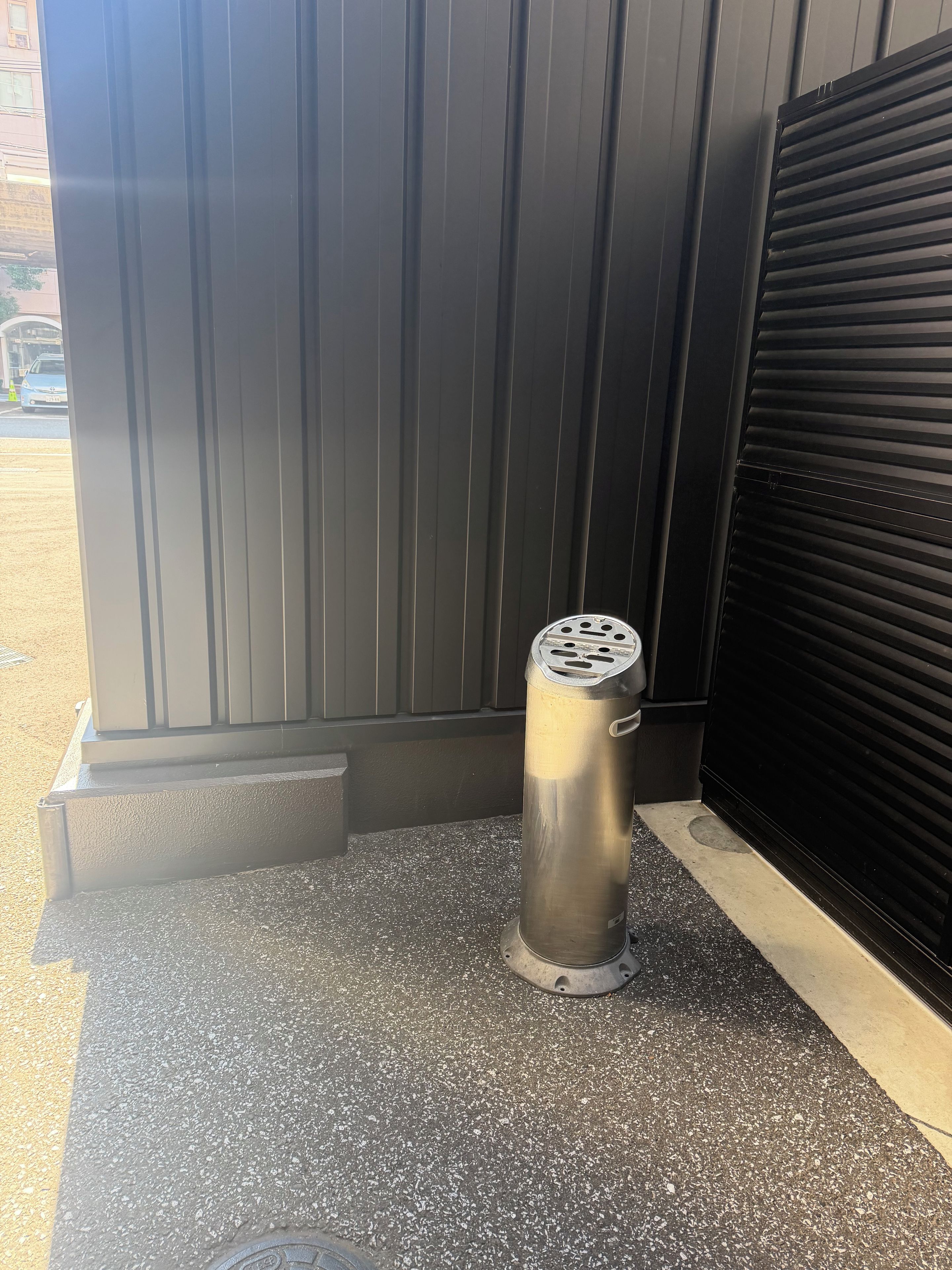 A metallic trash can with a circular top and a handle, placed on a textured pavement beside a closed black shutter door.
