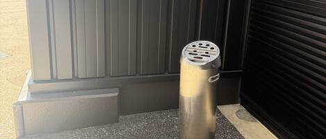 A metallic trash can with a circular top and a handle, placed on a textured pavement beside a closed black shutter door.