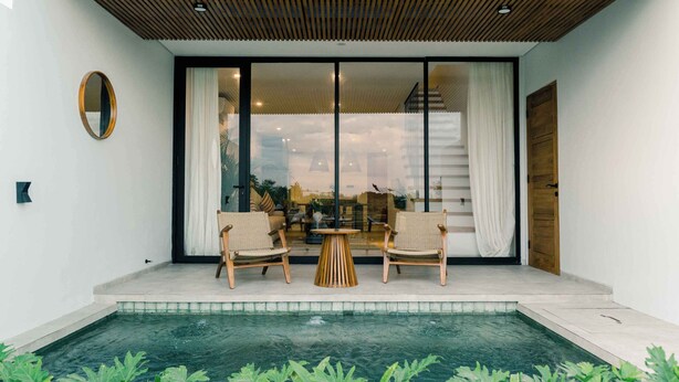 A modern outdoor patio with a pool, two wicker chairs, a small round table, and a wooden door. - The Menuai Bali Villas Canggu (Canggu)