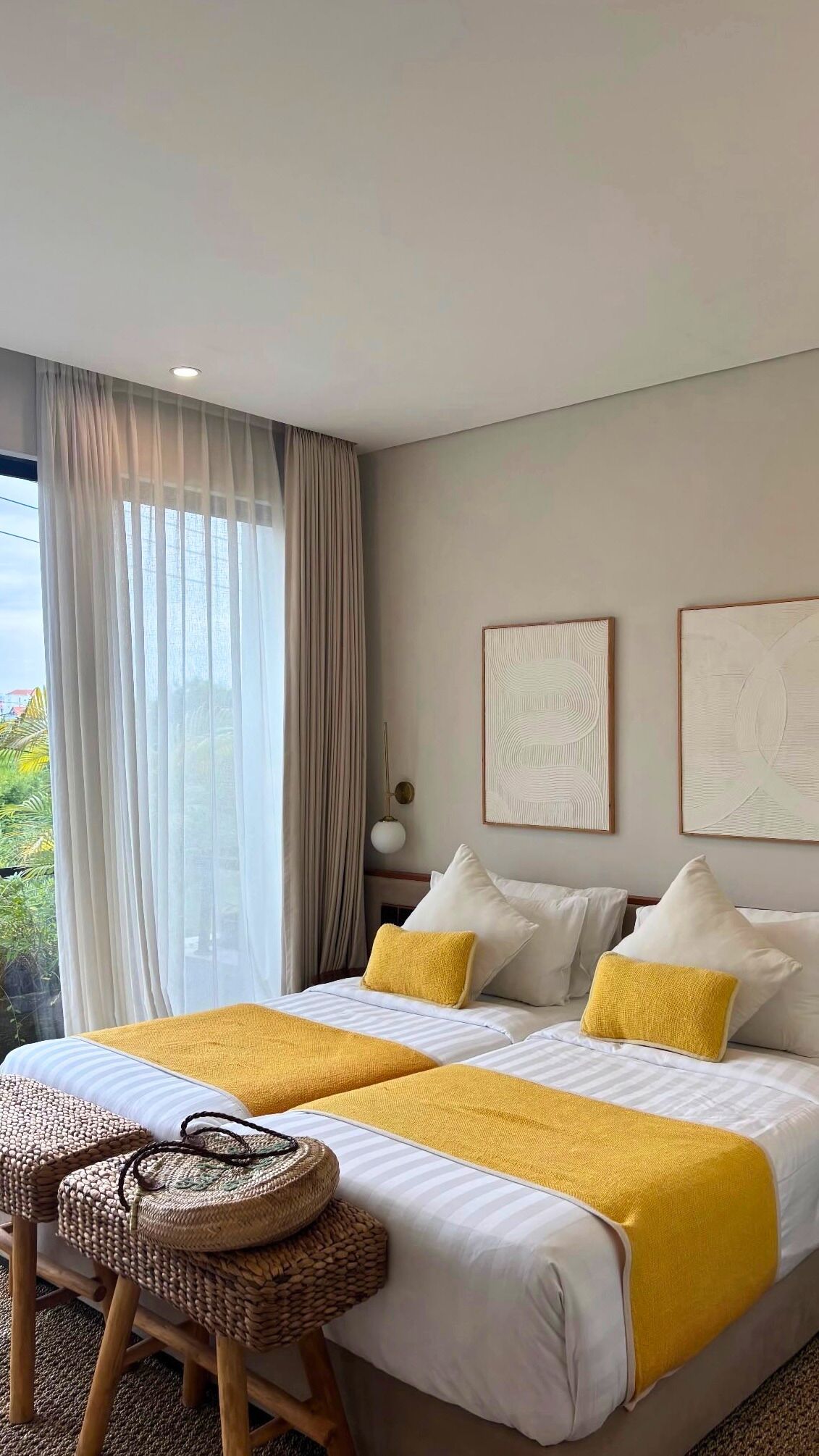 Una cama doble con ropa de cama blanca y amarilla, un taburete tejido y dos obras de arte colgadas en la pared.