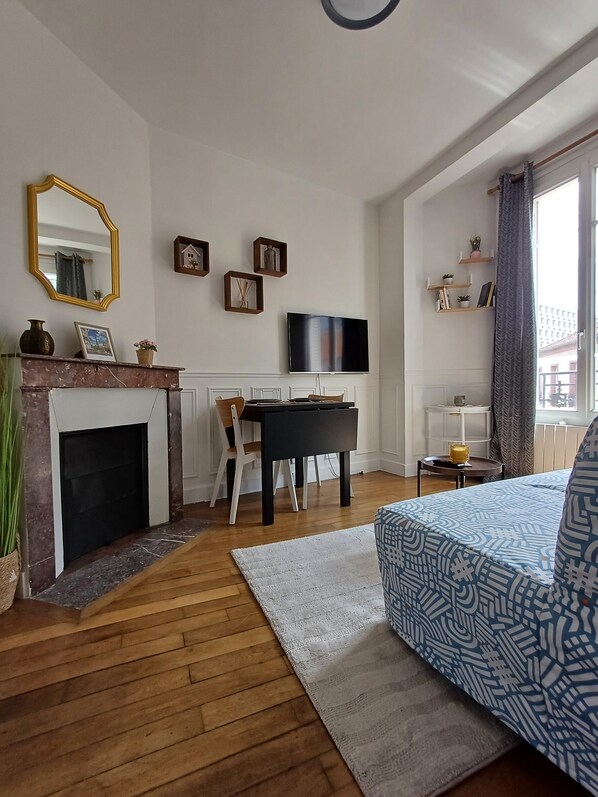 A modern living room with a fireplace, a dining area, a flat-screen TV mounted on the wall, and a bed with patterned bedding.