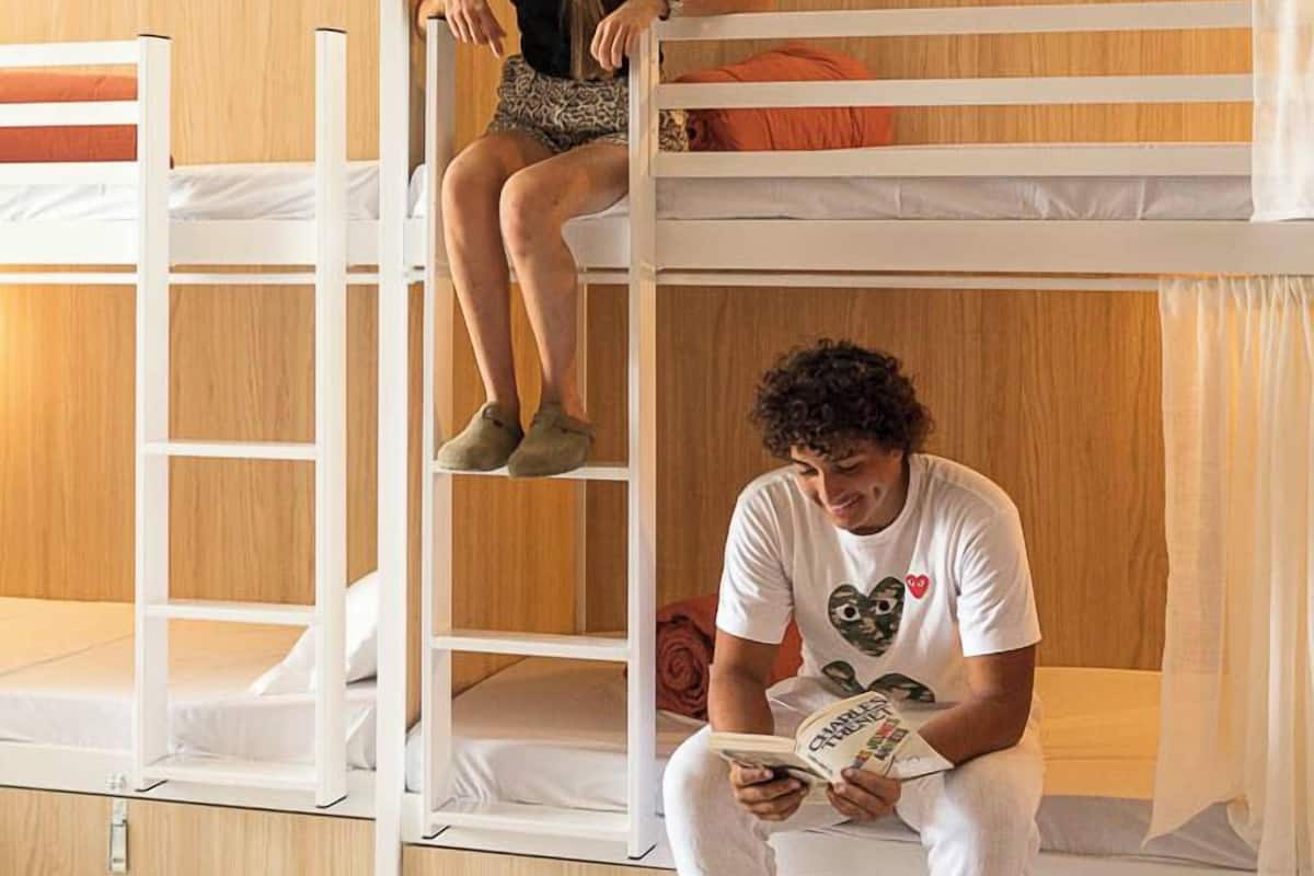 Ein Schlafraum mit Etagenbetten, einer Leiter, einer Person, die im oberen Bett sitzt, und einer weiteren Person, die unten ein Buch liest.