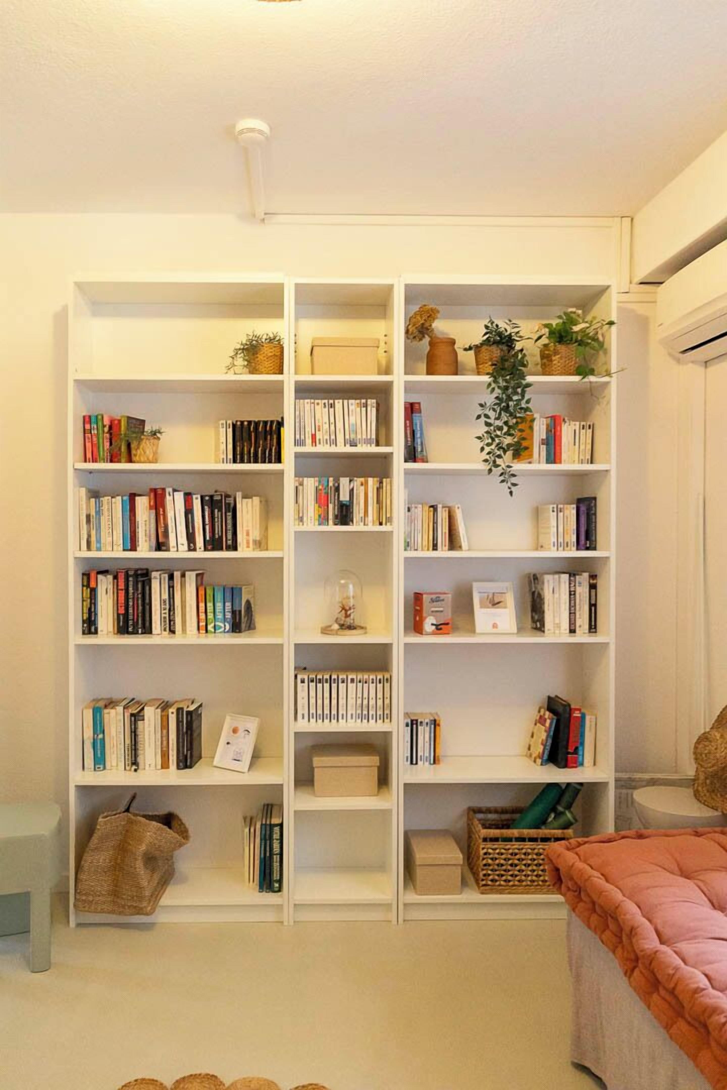 Une pièce avec une grande bibliothèque blanche remplie de livres et d’objets décoratifs, une petite table avec un panier, et une couverture orange douillette sur le sol.