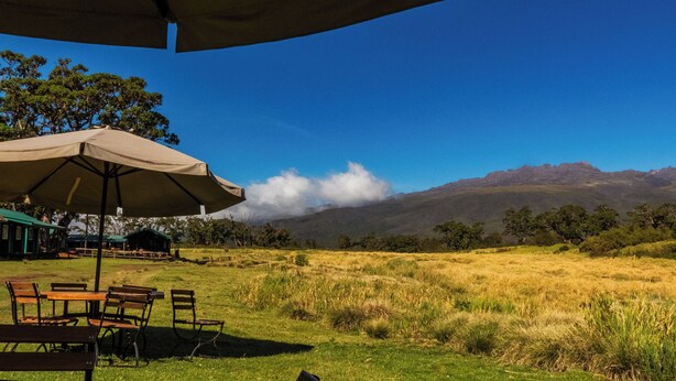 A grassy field with a table and chairs under a large umbrella, overlooking a mountain range. - Anabas Mount Kenya Lodge (Igoji)