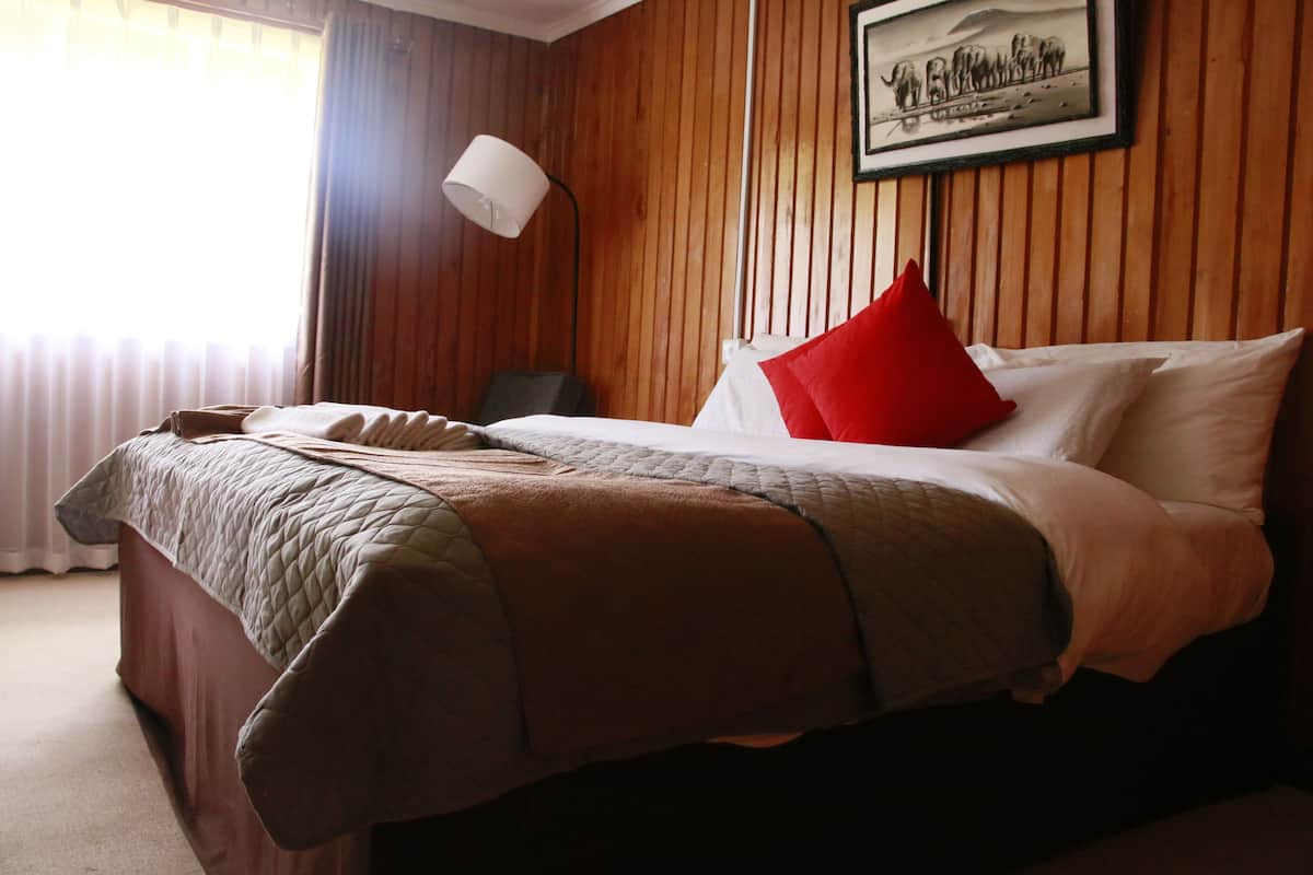 Et hotelværelse med en væg af træpaneler, en seng med hvide og røde puder, et quiltet dyne og en sengetavlelampe samt et billede i ramme på væggen.