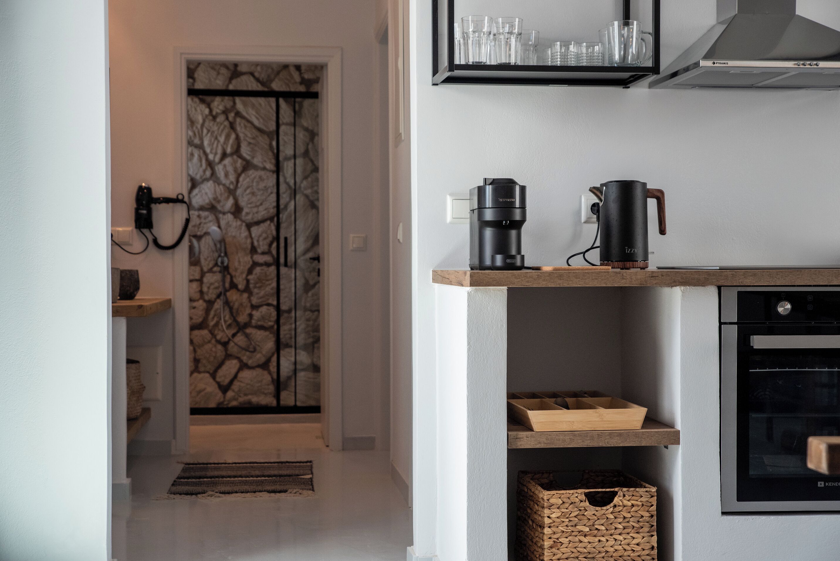 Et moderne køkken med indbygget kaffemaskine, elkedel og Keurig-kaffemaskine på en træbordplade. En dør med stengulvmønster fører til et andet rum, og der er en kurv på gulvet.