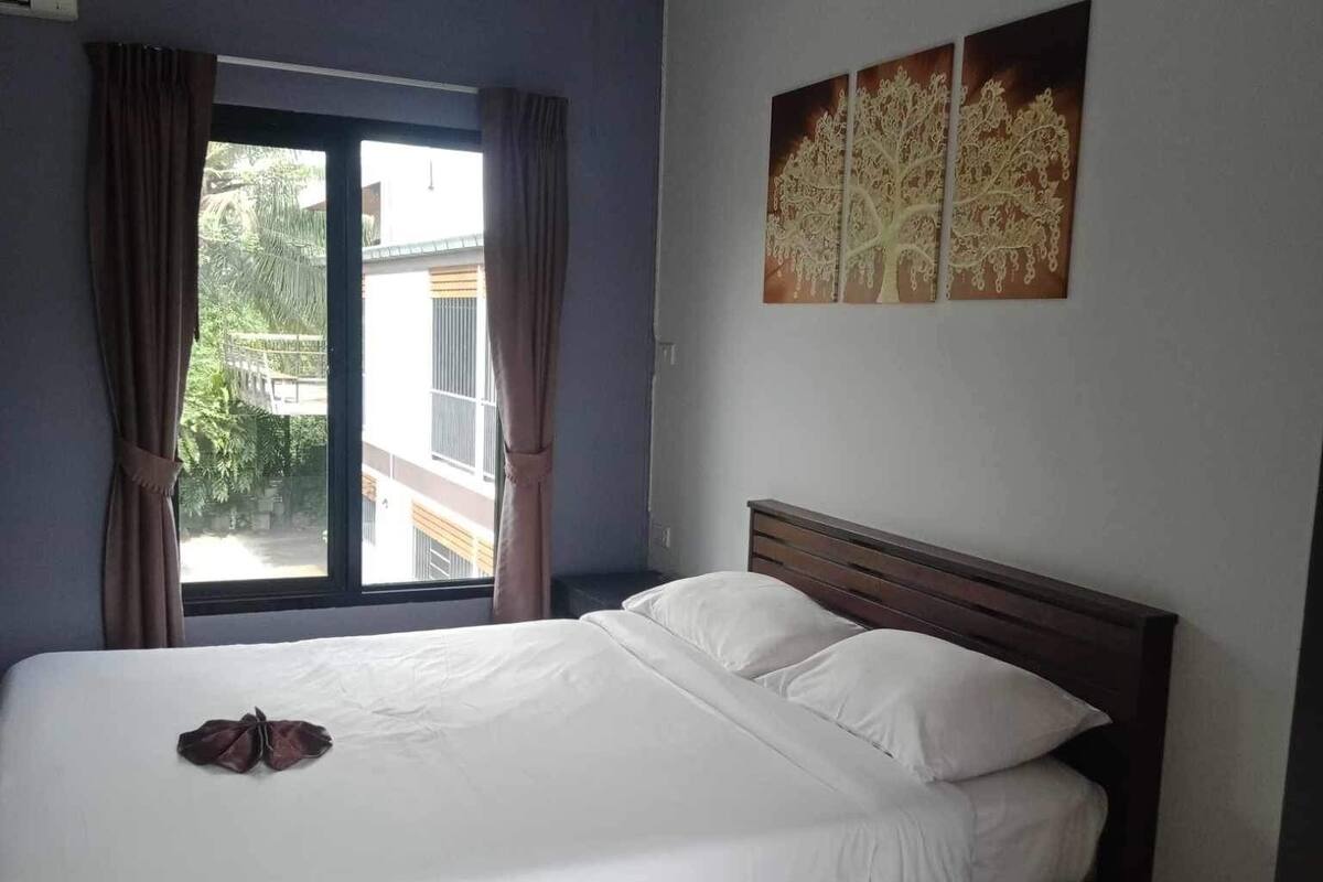 A bedroom with a large bed, a window with curtains, a wall-mounted artwork, and a ceiling vent.