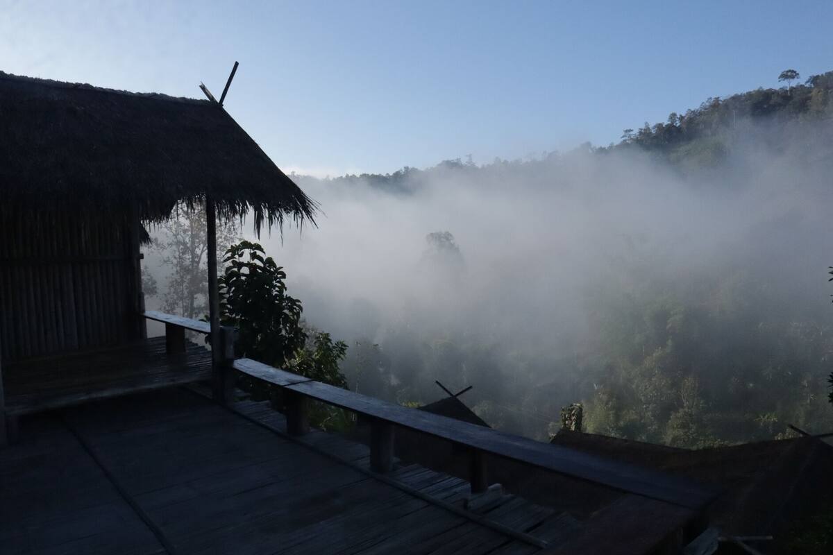 A wooden deck with a thatched-roof structure, overlooking a misty forested valley.
