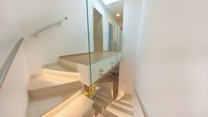 Un escalier avec des rampes et une main courante en verre, menant à un palier où se trouvent une porte et une petite table sur laquelle sont posés divers objets.
