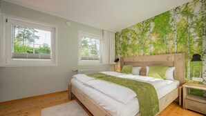 Un dormitorio con una cama de madera, dos ventanas, un papel tapiz con motivos de bosque verde y una mesita de noche con lámpara.