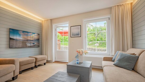 Una sala de estar moderna con un sofá color beige, dos otomanas grises, un televisor de pantalla plana en la pared y una ventana con vistas a follaje y un paraguas rojo en el exterior.