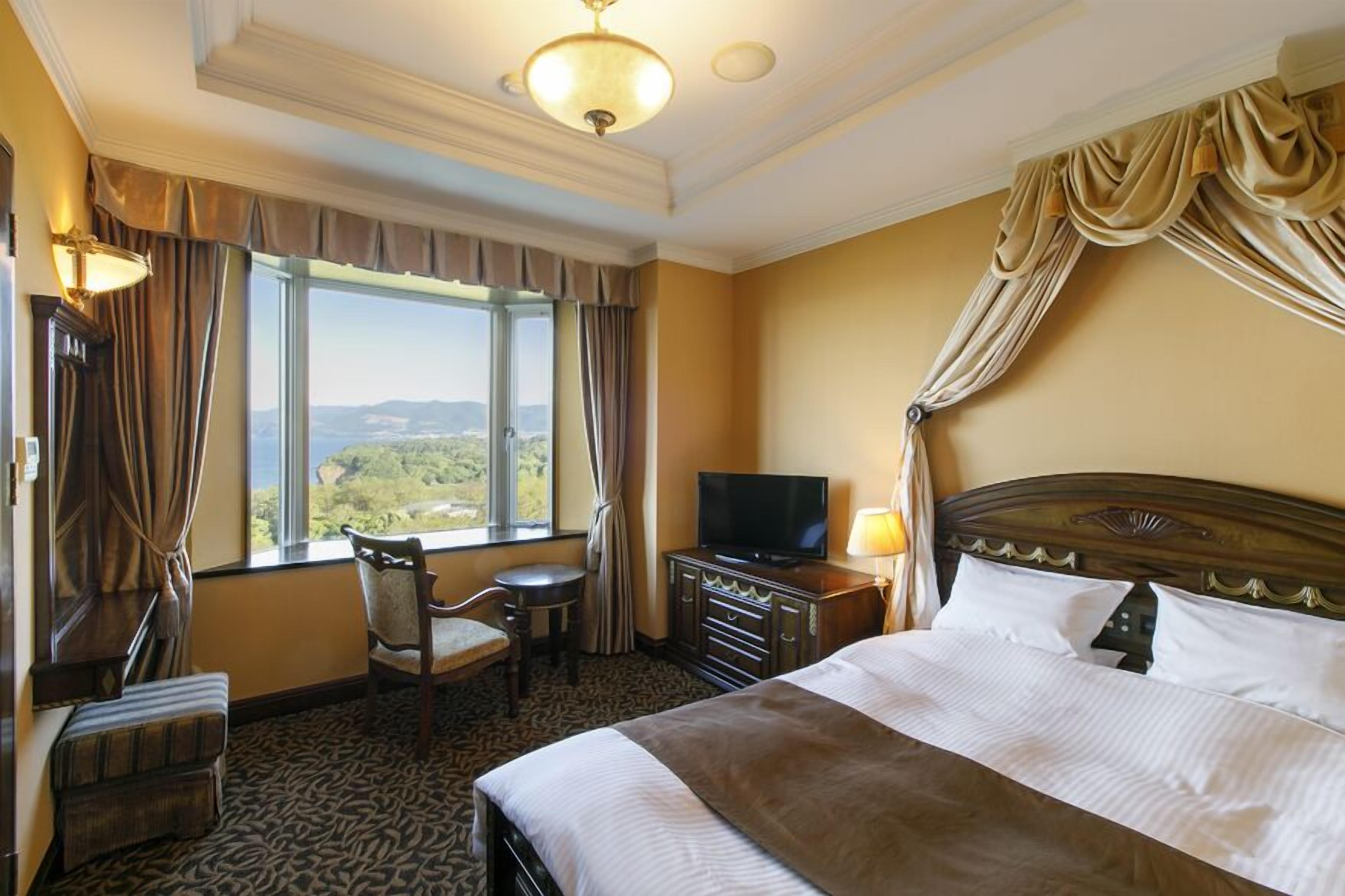 Een hotelkamer met een groot bed, een televisie, een bureau met stoel en uitzicht op het groen door het raam.