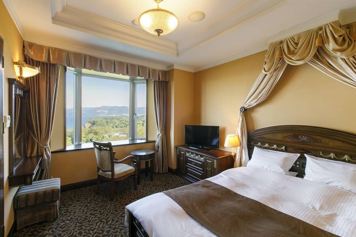 A hotel room with a large bed, a television, a desk with a chair, and a view of the outdoors through the window.