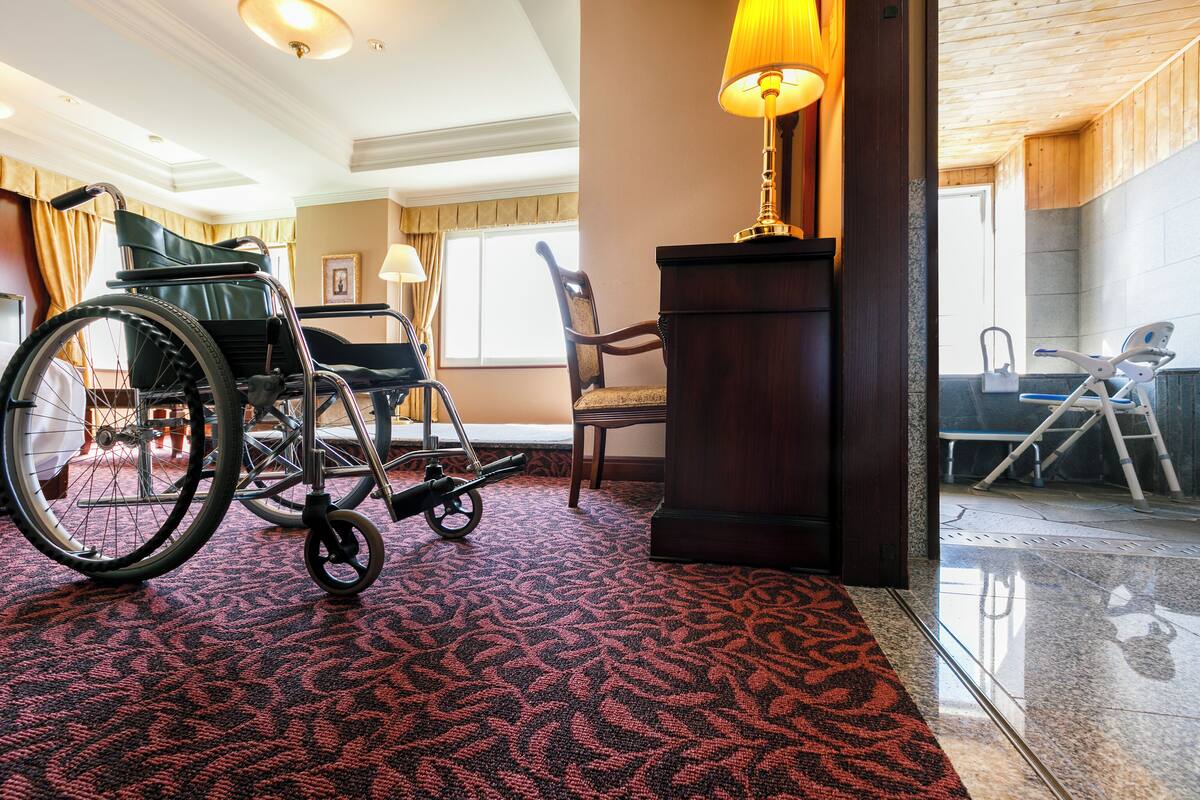 A wheelchair in a hotel lobby with a patterned carpet, a wooden chair, and a lamp.