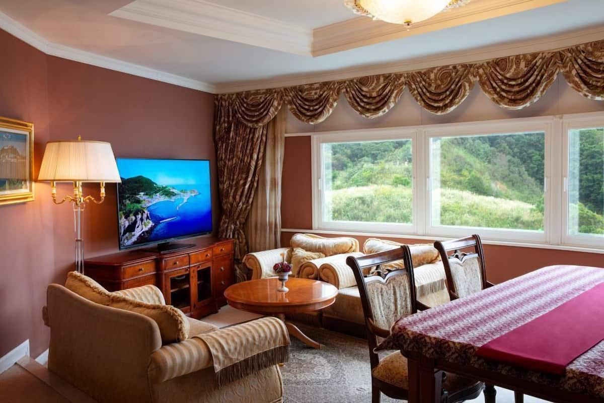 A living room with a television, sofa, dining table, and a view of greenery through large windows.