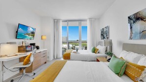 A modern hotel room with a large bed, a desk with a computer, a TV, and a view of the city through large windows. - Amazing Studio- Bay View & Amenities (Hallandale Beach)
