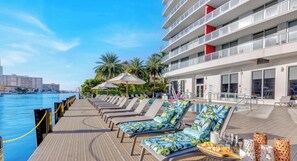 A poolside area with lounge chairs and umbrellas, a waterfront view, and a modern building with balconies.