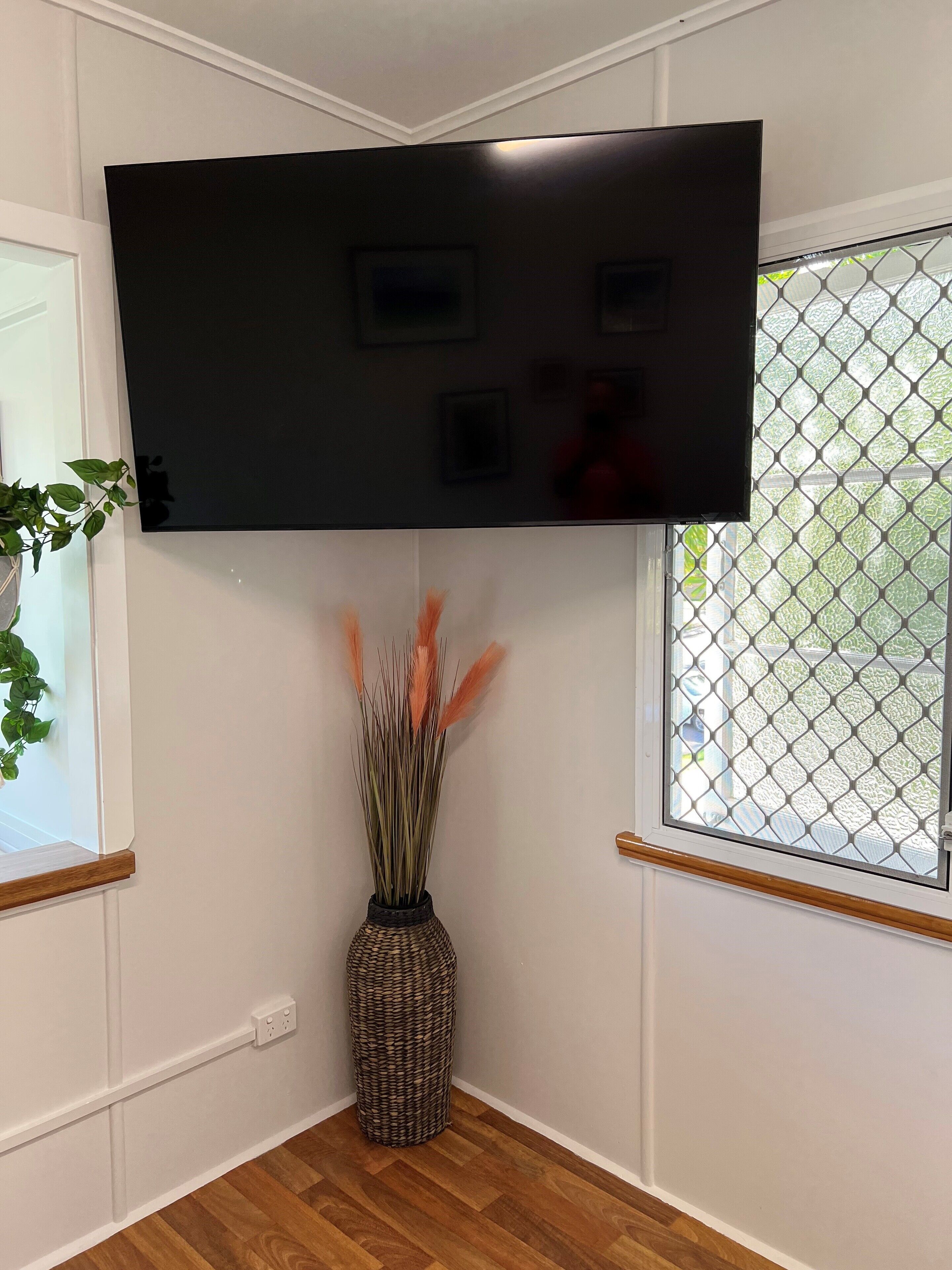 A room with a mounted flat-screen TV, a woven basket with decorative plants, and a window with a metal grid.