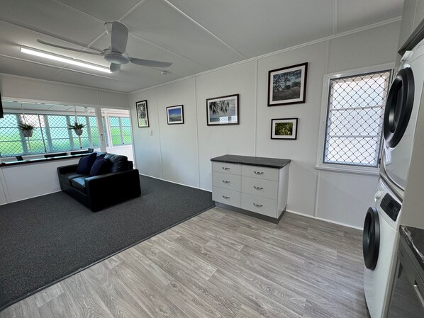 A modern laundry room with a large washing machine, a grey carpet, a black leather sofa, and a window with a view of greenery. - Essex Court (North Ward)