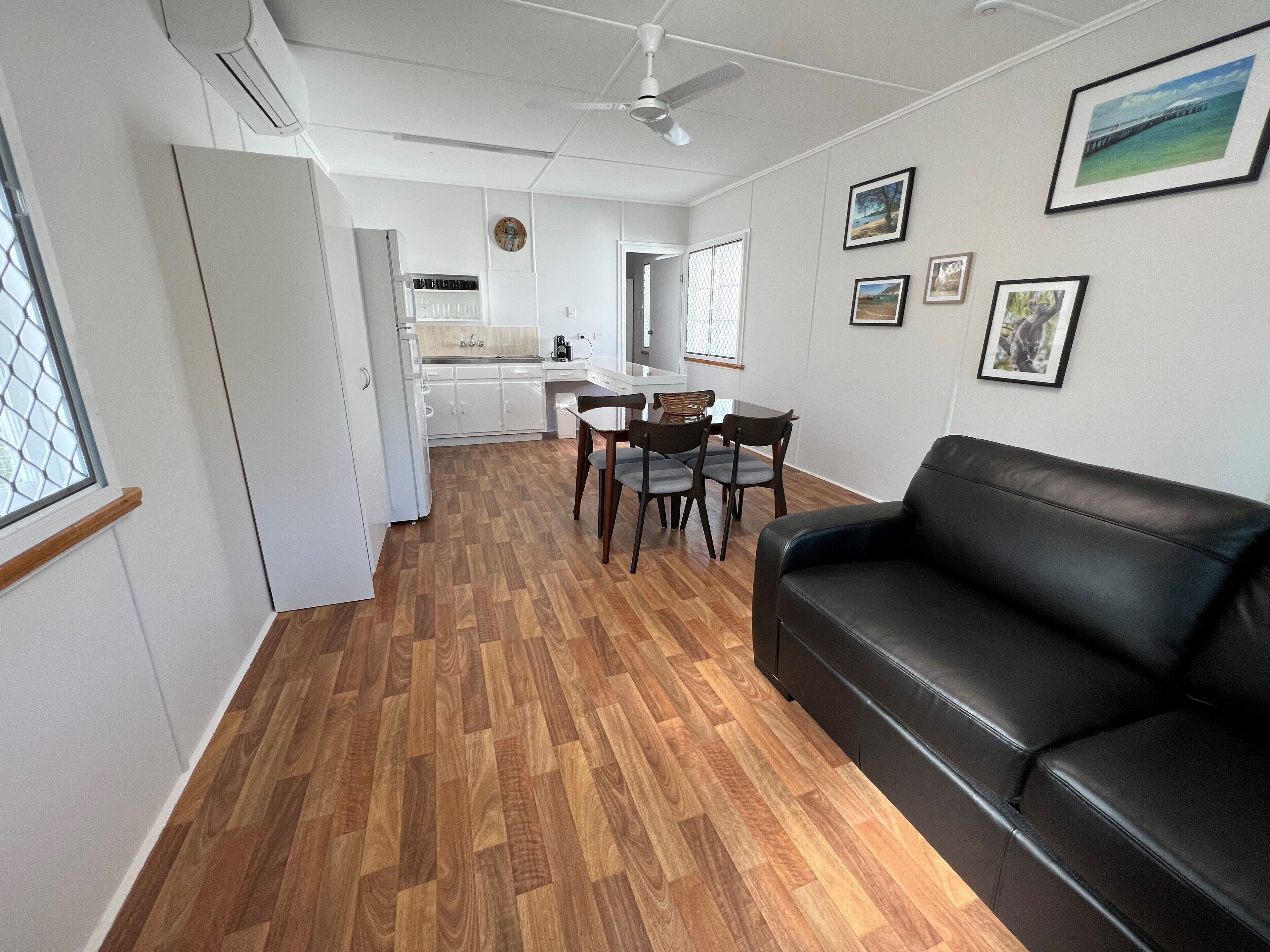 Una sala de estar moderna con un sofá de cuero negro, piso de madera, un comedor con mesa y sillas, y una cocina al fondo.