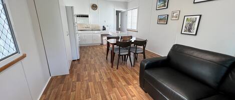Una sala de estar moderna con un sofá de cuero negro, piso de madera, un comedor con mesa y sillas, y una cocina al fondo.