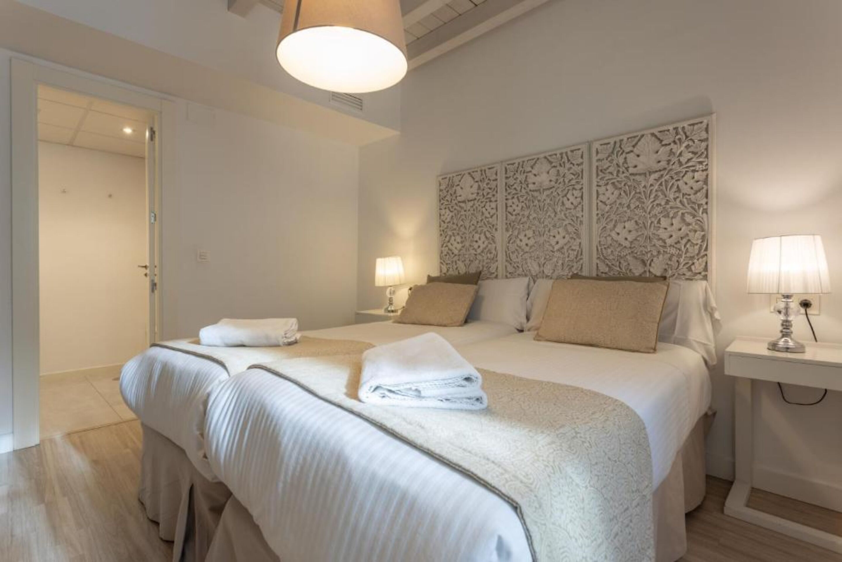 A hotel room with a large bed, two bedside tables with lamps, a decorative headboard, and a door leading to another room.