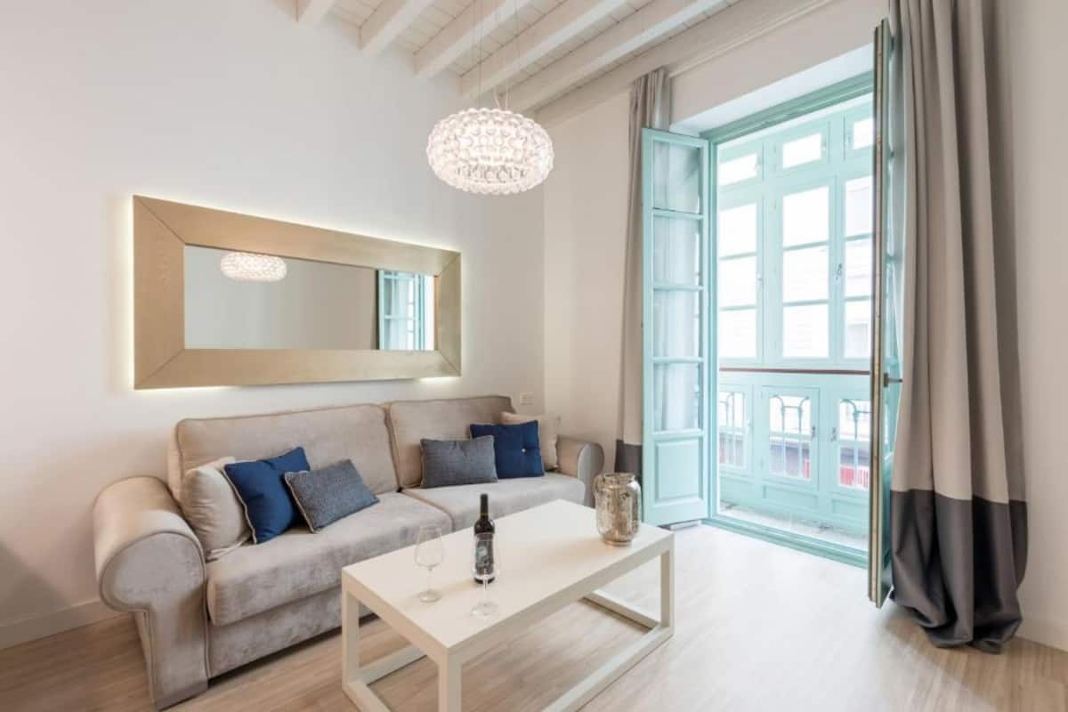 A modern living room with a sofa, a coffee table, a chandelier, and a large window with curtains.