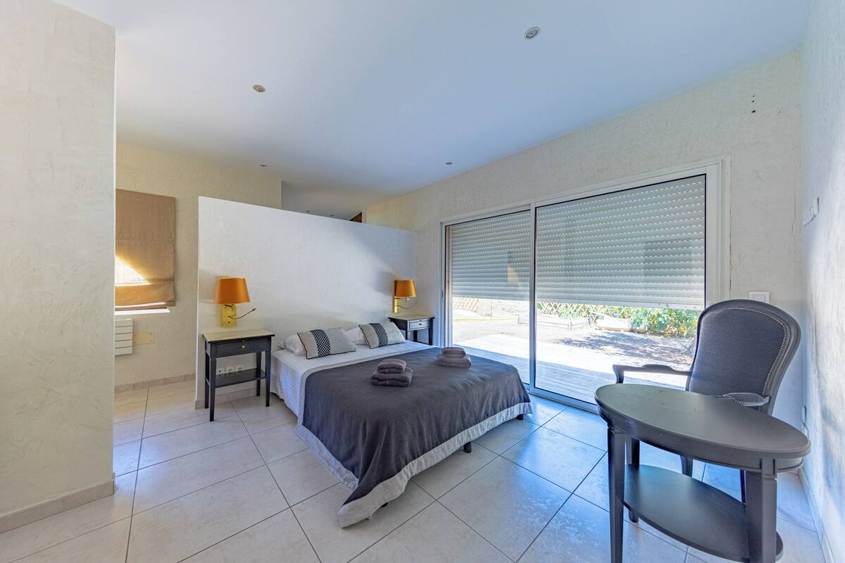Une chambre à coucher comprenant un lit, des tables de chevet, une chaise et une grande fenêtre avec des stores.