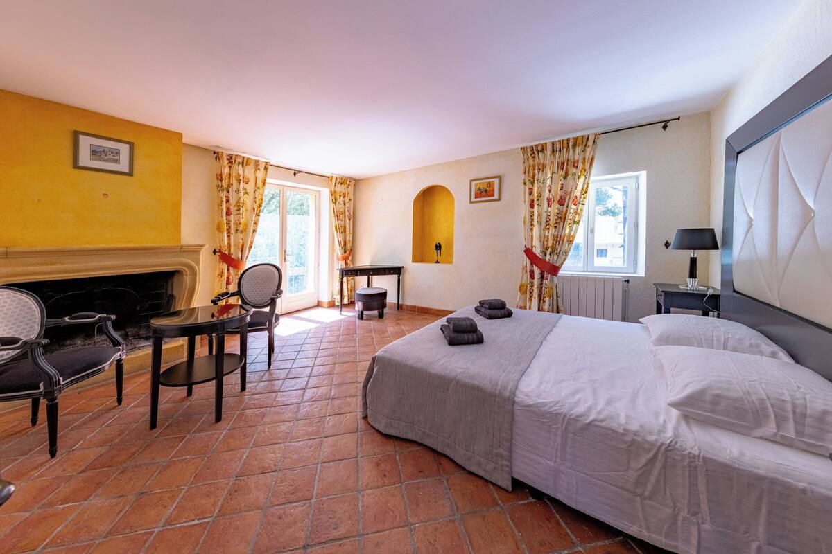 Une chambre d’hôtel avec un lit, une chaise, une petite table, une cheminée et une fenêtre avec des rideaux.