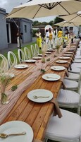 Una larga mesa de madera dispuesta con platos blancos y cucharas doradas, rodeada de sillas blancas, bajo grandes sombrillas beige.