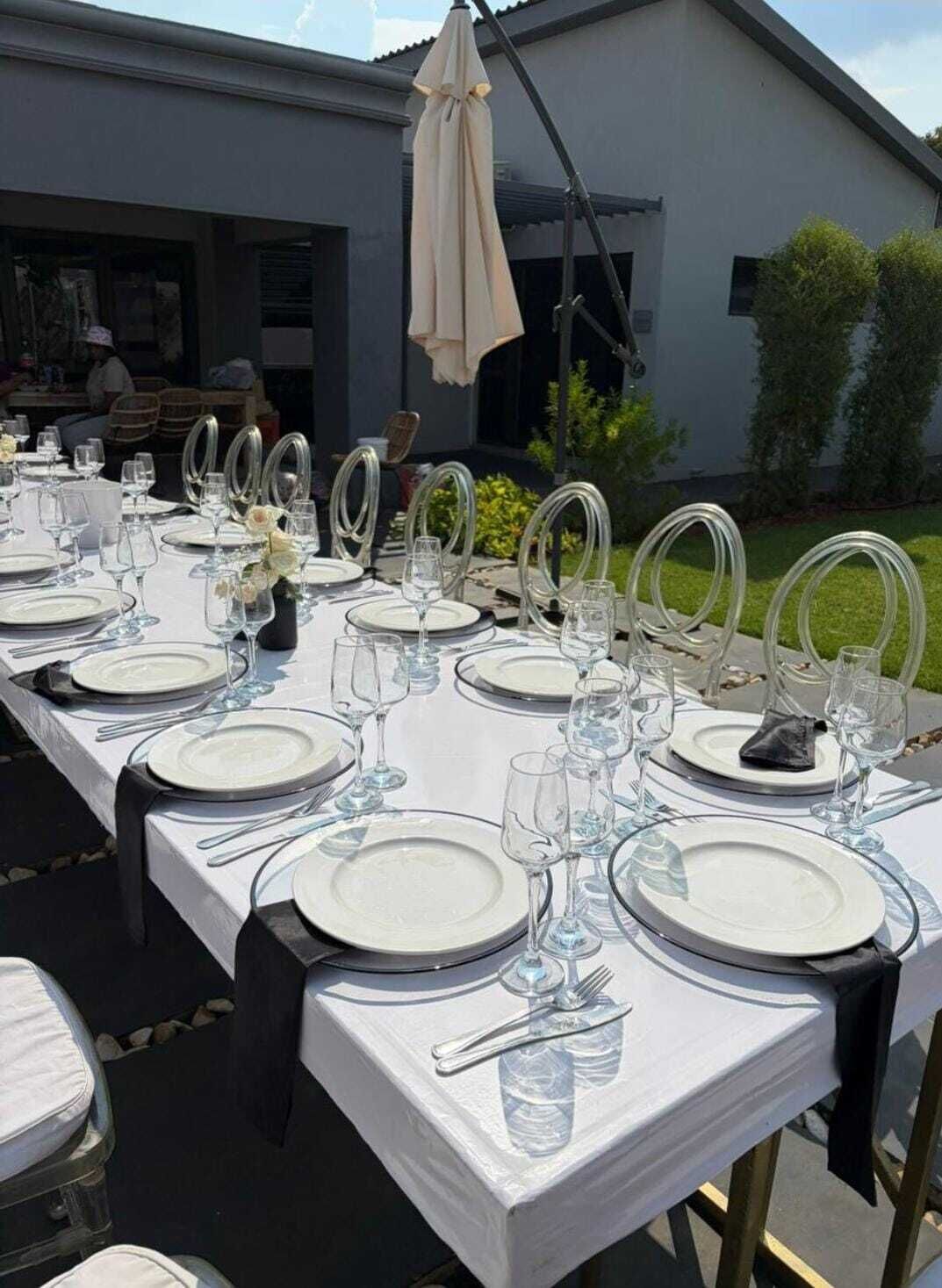 A long outdoor dining table set with white tablecloths, clear glassware, and metal chairs.
